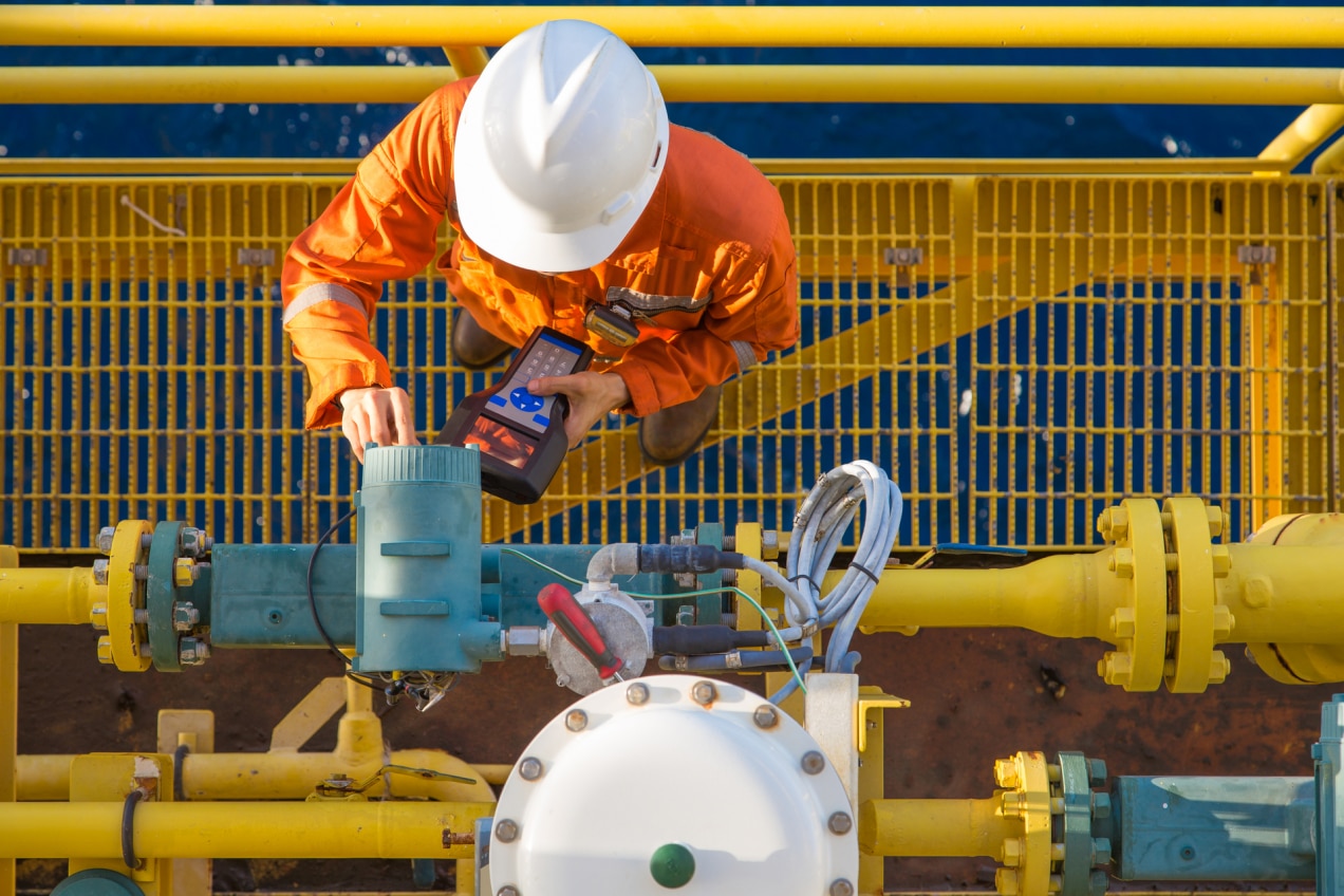 A technician in an orange safety suit and white hard hat is performing maintenance or diagnostic checks on industrial equipment. The setting includes yellow piping and various valves, with the technician using a handheld device to interact with the equipment. The scene is likely set in an industrial environment, such as an oil rig, refinery, or chemical plant, emphasizing safety and precision in operations.