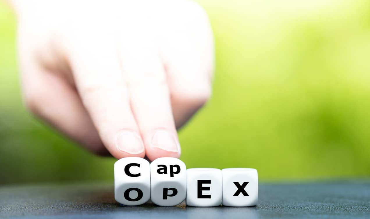 The image shows a hand adjusting dice to display the words "CapEx" and "OpEx." The dice are arranged in a way that highlights the transition between the two terms. "CapEx" stands for Capital Expenditure, and "OpEx" stands for Operational Expenditure. The background is blurred with a gradient of light green, providing a soft and focused view on the dice and hand. This visual represents the financial concepts of capital and operational expenditures in a business context.