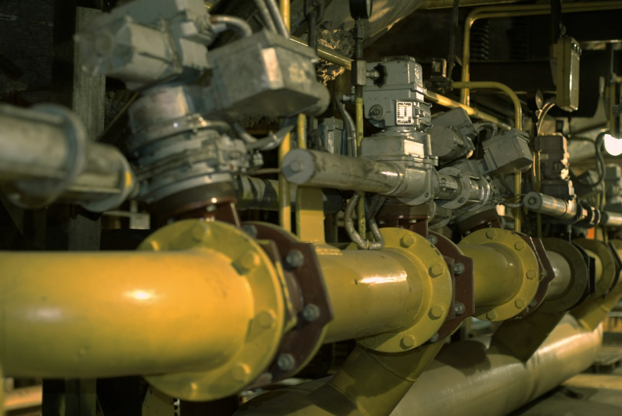 The image shows a close-up view of a series of industrial valves and pipelines within a processing plant. The yellow pipes are interconnected with several control valves, indicating a complex fluid or gas distribution system. The equipment appears well-used, with signs of wear and some buildup, suggesting an operational environment focused on control and maintenance of process flows.