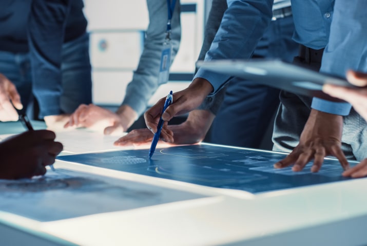 A team of professionals is gathered around a large, illuminated table, engaged in a collaborative discussion. They are examining blueprints and documents spread across the table, with some individuals pointing and using pens to highlight key areas. The setting suggests a high-tech, innovative environment, likely focused on engineering, design, or project planning. The atmosphere conveys teamwork, precision, and the dynamic process of developing complex solutions.