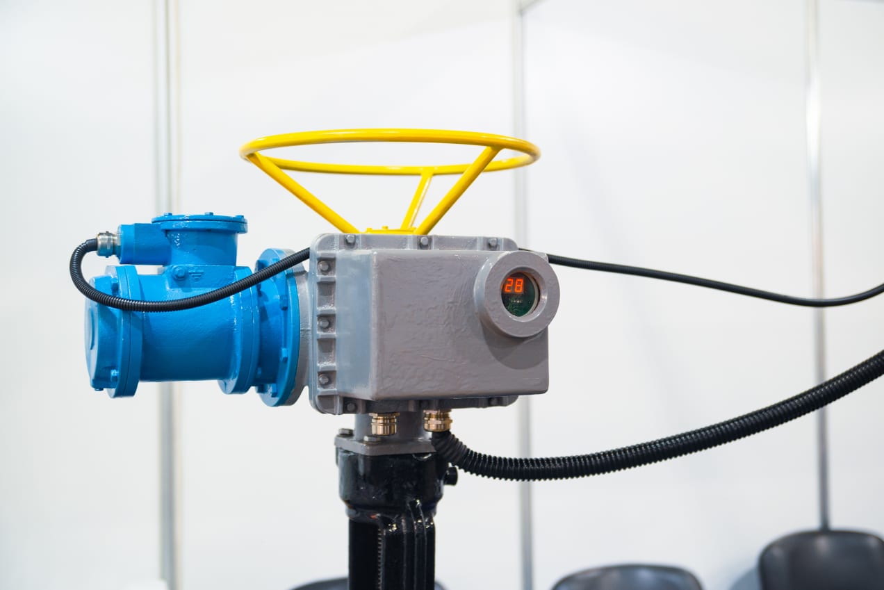 The image shows a close-up of an industrial actuator with a bright yellow handwheel and blue housing. The actuator is mounted on a piece of industrial equipment and features a digital display showing the number "28". The setup appears to be part of a system that controls the flow or pressure within an industrial process, highlighting the equipment's robust and technical design.