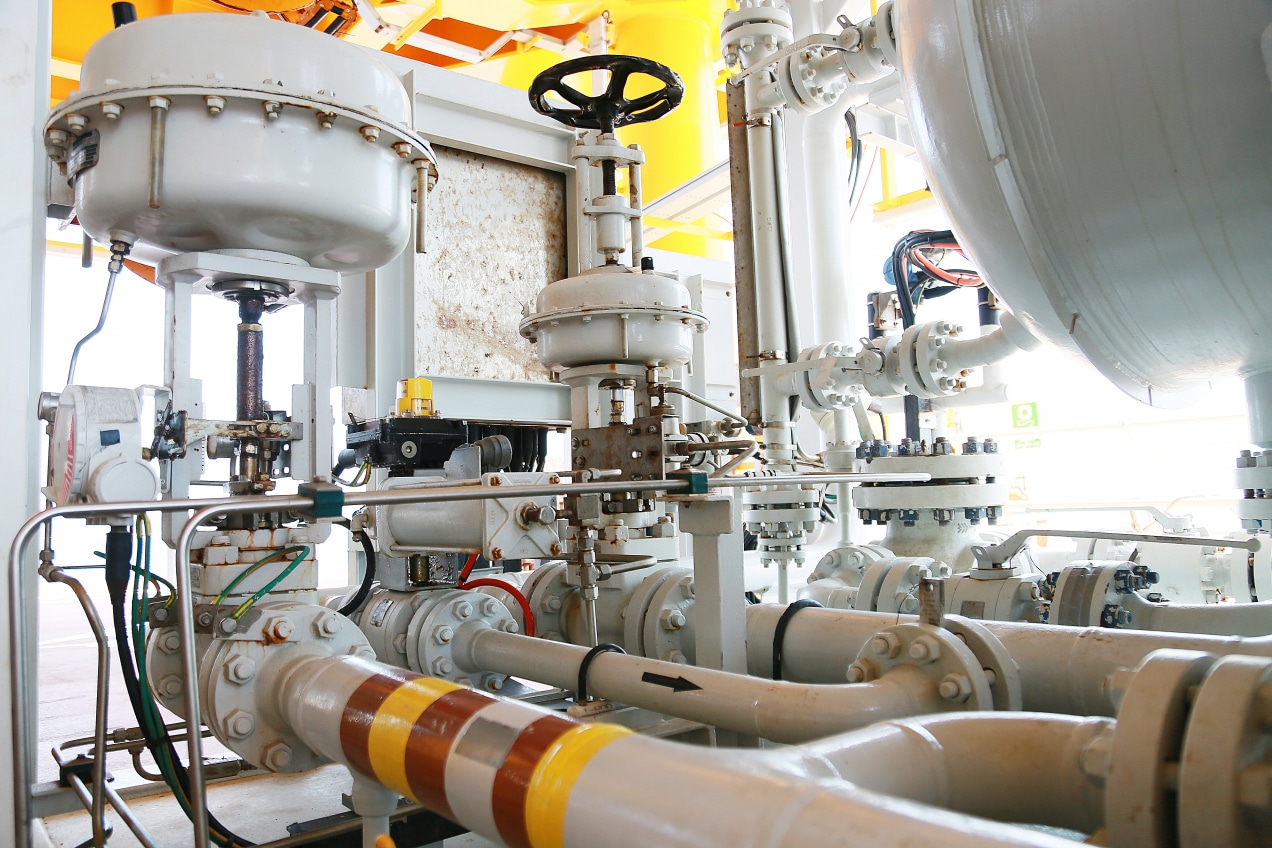 The image depicts a complex industrial setup featuring various valves, pipes, and equipment, likely within a processing or manufacturing plant. The scene includes: Valves: Prominently visible are several large control valves with handwheels for manual operation. Pipes: Numerous interconnected pipes of different sizes, some labeled with directional arrows and color-coded bands for identification. Instrumentation: Gauges, sensors, and other control instruments are attached to the valves and pipes for monitoring and controlling the process. Actuators: Several pneumatic or hydraulic actuators connected to the valves for automated control. Background: The equipment is situated on a platform with a yellow structural framework, indicating an industrial environment, possibly in oil, gas, or chemical processing.