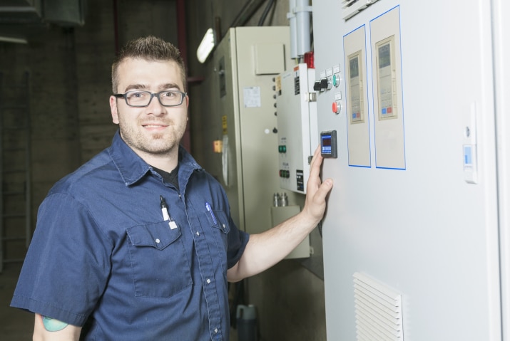 A technician wearing glasses and a dark blue work shirt is standing next to an industrial control panel. He is smiling and has one hand on the panel, which features several digital displays and control buttons. The background shows other industrial equipment and control boxes, indicating a setting within a manufacturing or processing facility. The technician's demeanor suggests confidence and expertise in managing and operating the equipment.