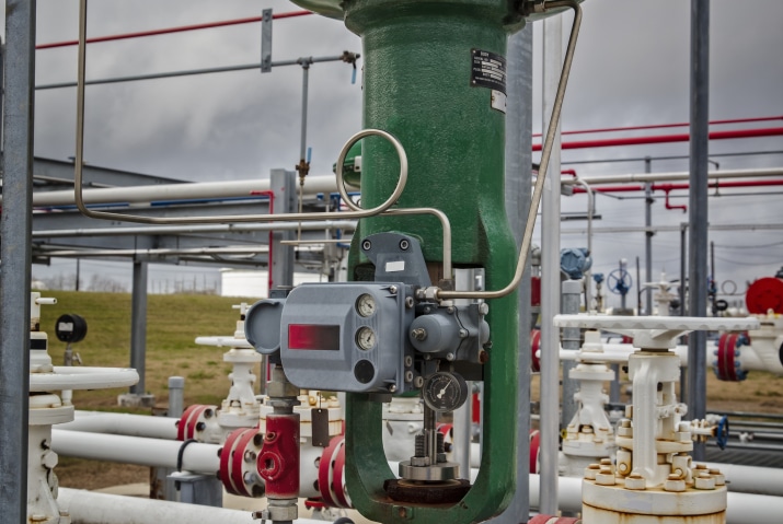 The image shows an industrial control valve in a field setting, likely part of a larger piping system. The valve body is green, with attached instrumentation, including a grey actuator or positioner with gauges and control lines connected. The surrounding area features various pipes and additional valves, indicating a complex network of fluid control. The background includes a grassy area and overcast sky, emphasizing the outdoor environment of the industrial site.