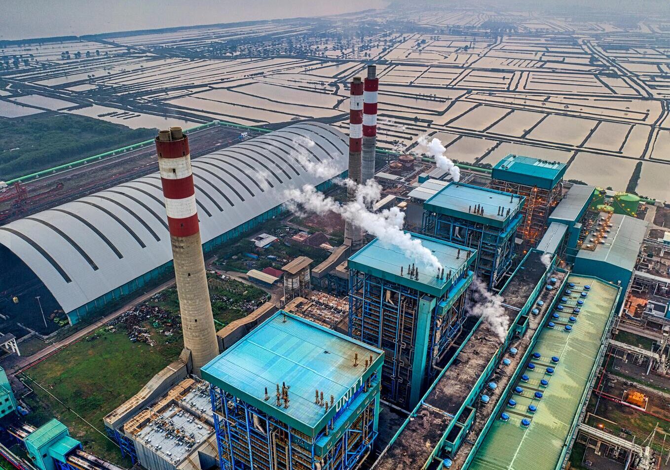 Aerial view of an industrial facility featuring large chimneys emitting steam or smoke, surrounded by extensive infrastructure. The facility has multiple blue and turquoise buildings with rooftop equipment, and the landscape beyond the facility includes a vast area of agricultural fields or salt pans, stretching towards the horizon under a hazy sky.