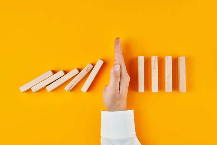 The image depicts a hand interrupting a sequence of falling wooden dominoes against a bright yellow background. The dominoes on the left side of the hand have fallen, while those on the right remain standing. The hand, positioned vertically with fingers together, acts as a barrier, stopping the chain reaction of the falling dominoes. The image symbolizes the concept of intervention, prevention, or breaking a chain of events.