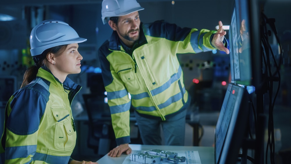 Two industrial engineers in high-visibility jackets and helmets are working together in a control room. They are closely monitoring data on multiple screens, with one engineer pointing at the display while discussing the information. The scene is set in a modern, dimly lit environment, indicating a focus on precision and advanced technology.