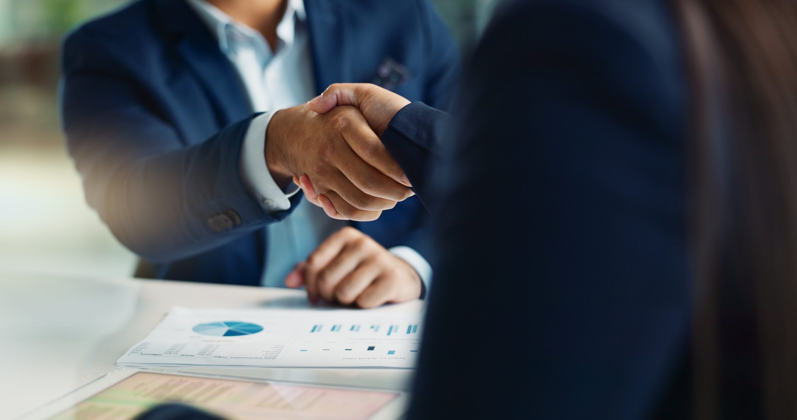 The image depicts two people in business attire shaking hands across a table. The focus is on their hands, suggesting a business agreement or partnership. A document with charts and graphs is partially visible on the table, indicating a formal business setting or negotiation. The background is blurred, keeping the attention on the handshake as the central theme.