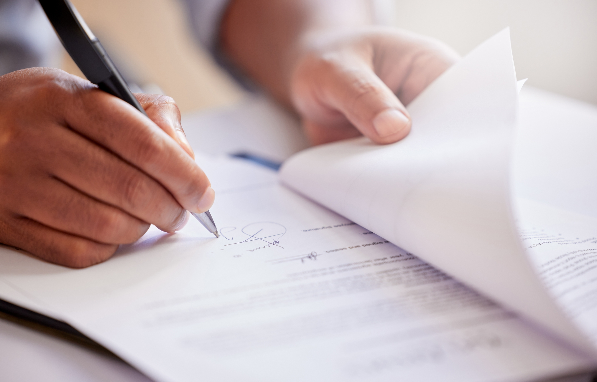 This image shows a close-up of a person's hands as they sign a document. The person is holding a pen and appears to be flipping through the pages while signing. The focus is on the action of signing, with the rest of the document slightly blurred in the background. The lighting is soft, highlighting the hand and pen, creating a professional and formal atmosphere.