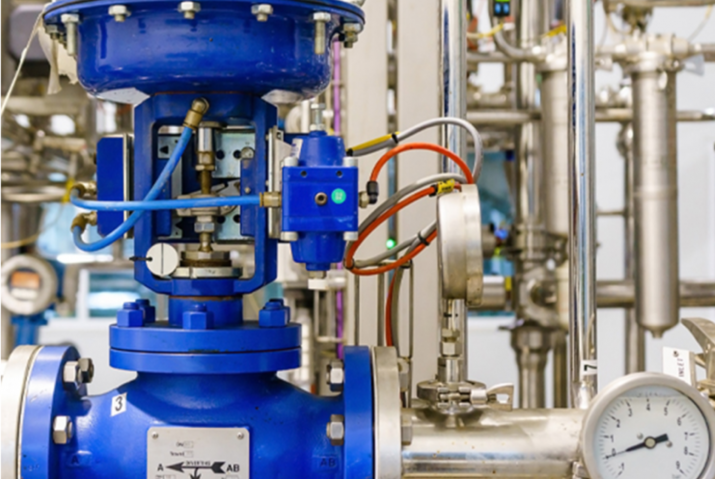 The image depicts a close-up view of a blue industrial valve installed within a complex piping system. The valve features various attached instruments, including gauges and control elements, with multiple wires and pipes connected to it. The background consists of stainless steel pipes and other industrial components, suggesting a setting such as a manufacturing plant or a processing facility.