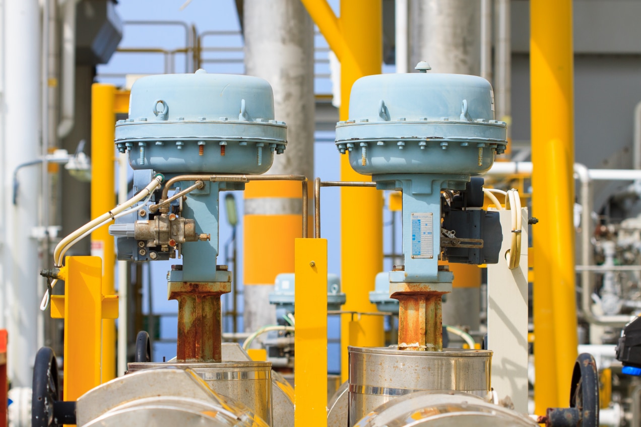 The image shows two control valves in an industrial setting, featuring light blue actuators mounted on top of yellow-rusted valve bodies. The valves are surrounded by vibrant yellow-painted structures, creating a contrast with the metallic components. In the background, various industrial equipment and pipelines are visible, contributing to the setting of a processing plant or refinery. The overall scene suggests an operational facility where valve performance and maintenance might be critical.