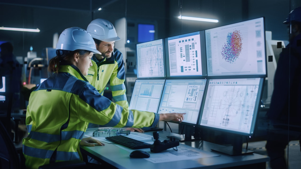 Two engineers in high-visibility jackets and hard hats monitor multiple data screens in an industrial control room, analyzing real-time information and system diagrams for maintenance and operational efficiency.