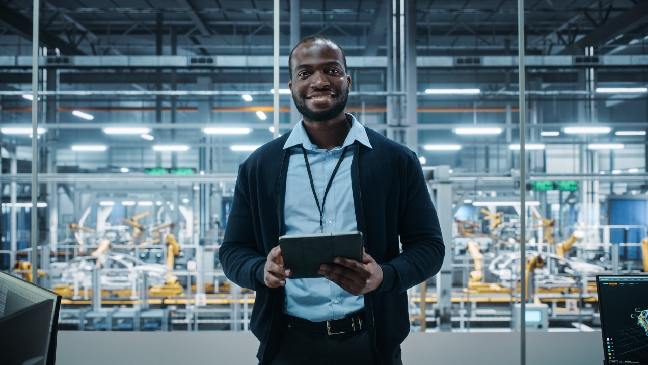 The image shows a man in a professional setting, standing confidently with a tablet in his hands. He is dressed in business-casual attire, wearing a light blue shirt and a dark cardigan, with an ID badge around his neck. Behind him is a modern industrial facility with advanced machinery, possibly an automated manufacturing or assembly line.