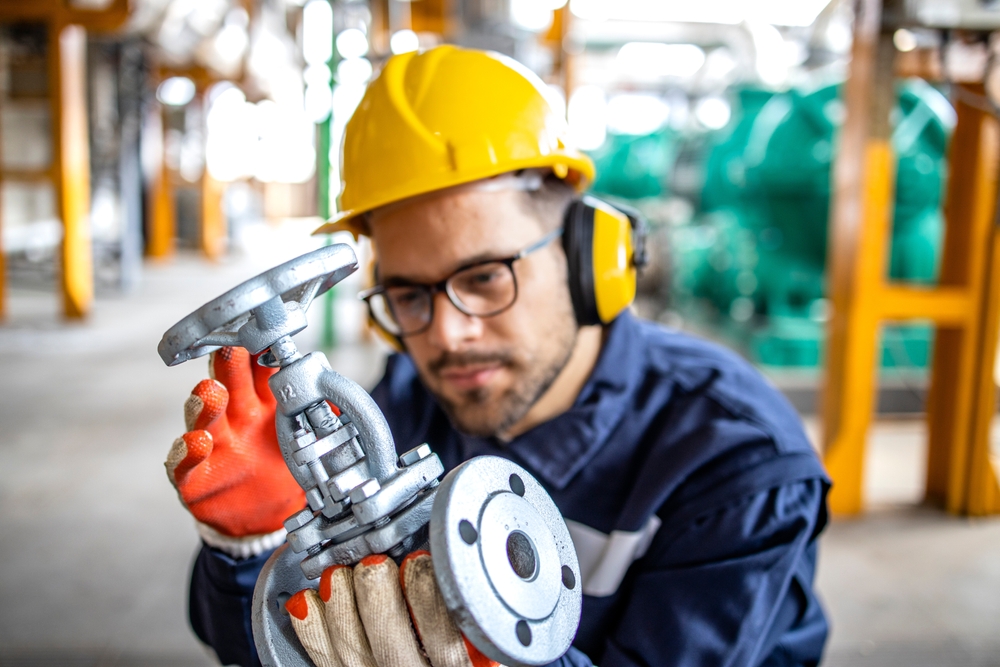 A worker in a safety helmet and ear protection inspects a metal control valve in an industrial setting. The worker is wearing safety gloves and appears to be focused on examining the valve's details. The background shows blurred industrial equipment, indicating a factory or plant environment.