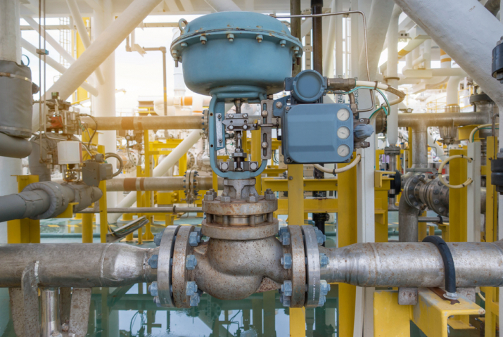 A large control valve installed in an industrial pipeline system, featuring a blue actuator on top. The valve is connected to surrounding pipes, which are part of a complex network within an industrial facility. The setup indicates the valve's role in regulating fluid flow through the system. Various support structures, pipes, and equipment are visible in the background, emphasizing the valve's importance in the industrial process.