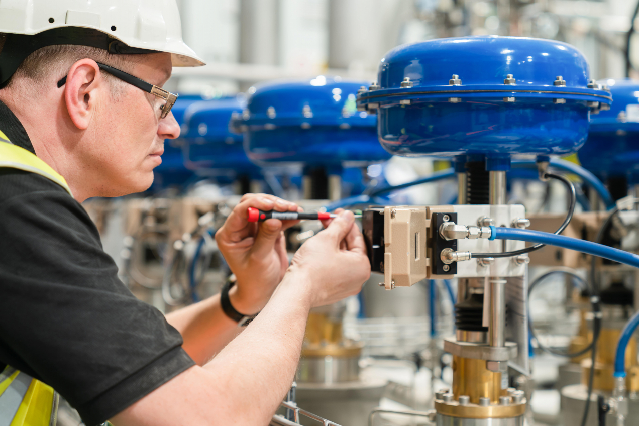 Engineer in a hard hat performing maintenance on a pneumatic control valve, using a screwdriver, with several blue valve actuators visible in the background.