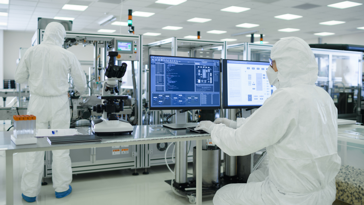 Two scientists in cleanroom suits working in a pharmaceutical manufacturing facility. One operates a computer with monitoring software, while the other attends to equipment in the background. A microscope and lab tools are visible on the desk, reflecting the highly controlled and sterile environment required for pharmaceutical production.