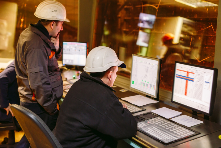 This image shows two industrial workers wearing blue coveralls, protective helmets, and safety glasses, collaborating in a factory or manufacturing setting.