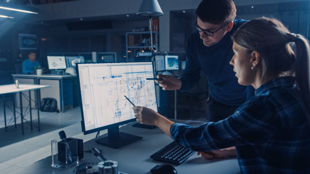 Two engineers working together in a modern office, analyzing technical blueprints on a computer monitor. One points at the screen with a pen while the other listens attentively, surrounded by other workstations and equipment.