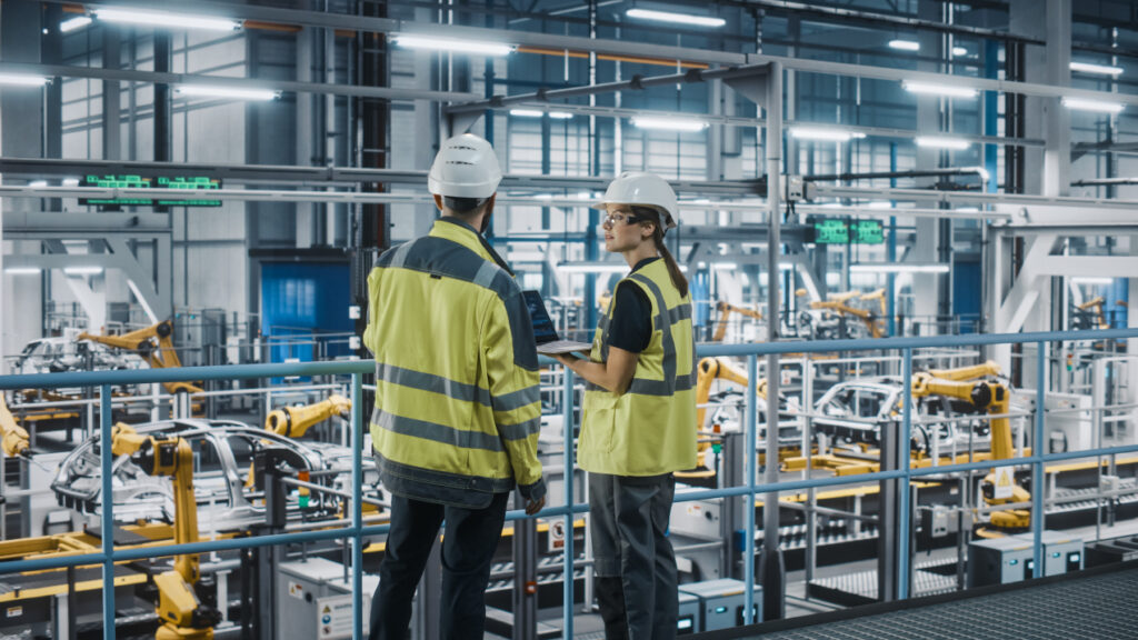 The image shows two industrial workers in high-visibility vests and hard hats standing on a metal platform overlooking a modern manufacturing facility. One of them is holding a laptop while they both observe the production process below. The factory floor is filled with automated machinery and robotic arms, likely involved in car manufacturing, as several car frames are visible. The scene is brightly lit, emphasizing the advanced, automated nature of the manufacturing environment.