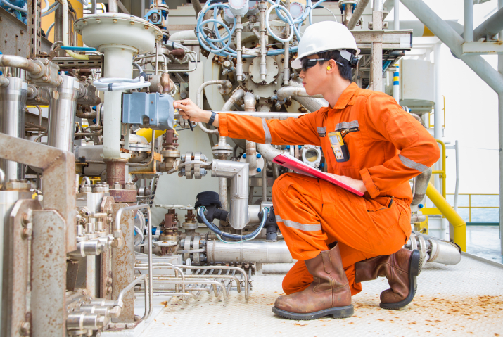 Engineer in orange protective gear and a hard hat performing maintenance checks on industrial equipment, holding a clipboard and adjusting a valve in a complex network of pipes and machinery.