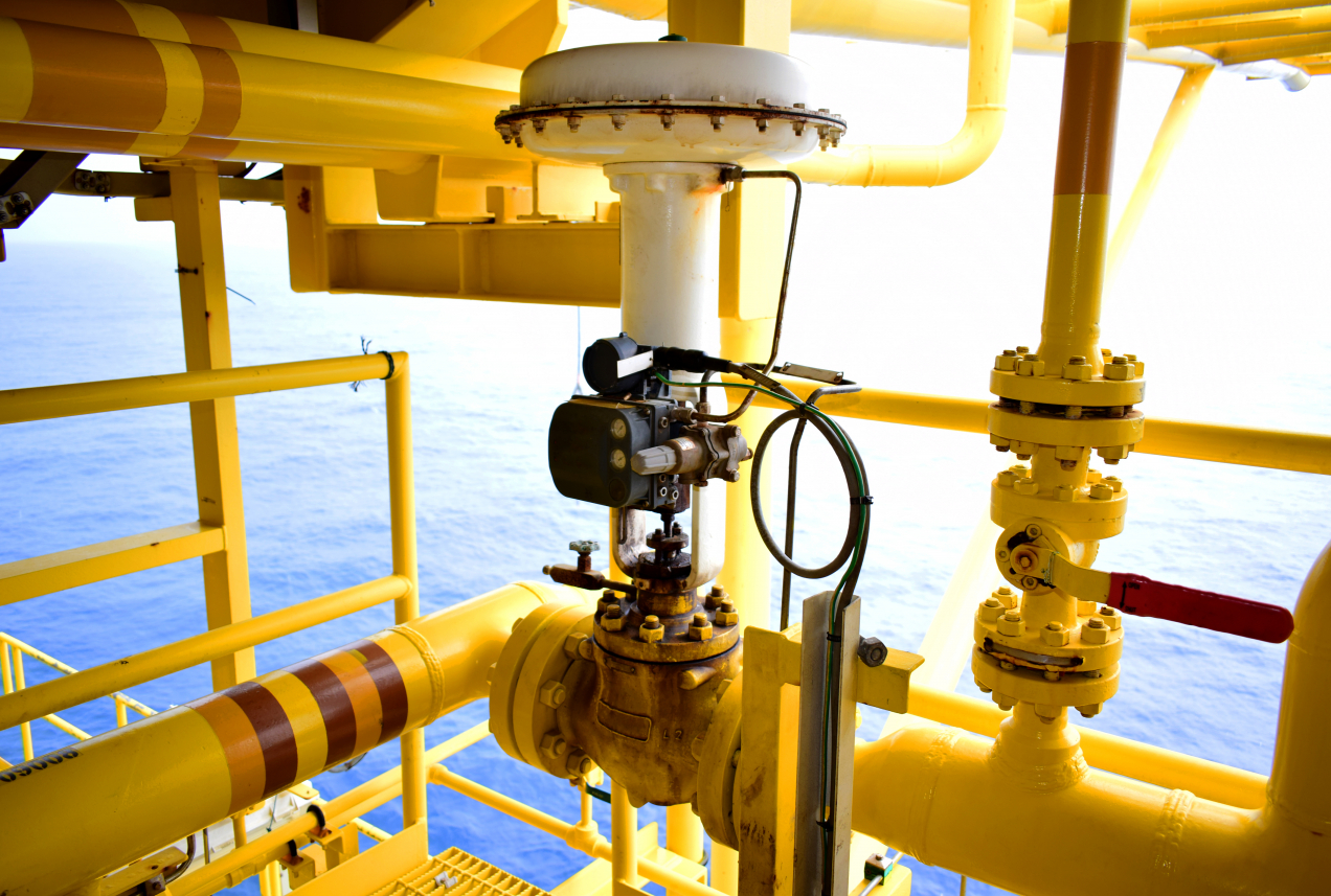 The image shows a close-up of a yellow industrial control valve installed on an offshore platform, with the ocean visible in the background. The valve is mounted on a series of yellow pipelines, and it is equipped with an actuator and a positioner, which are used to control the valve's operation. The setup is typical for oil and gas platforms, where precise control of the flow of fluids is critical. The bright yellow color of the equipment is a common safety measure to increase visibility in such environments.