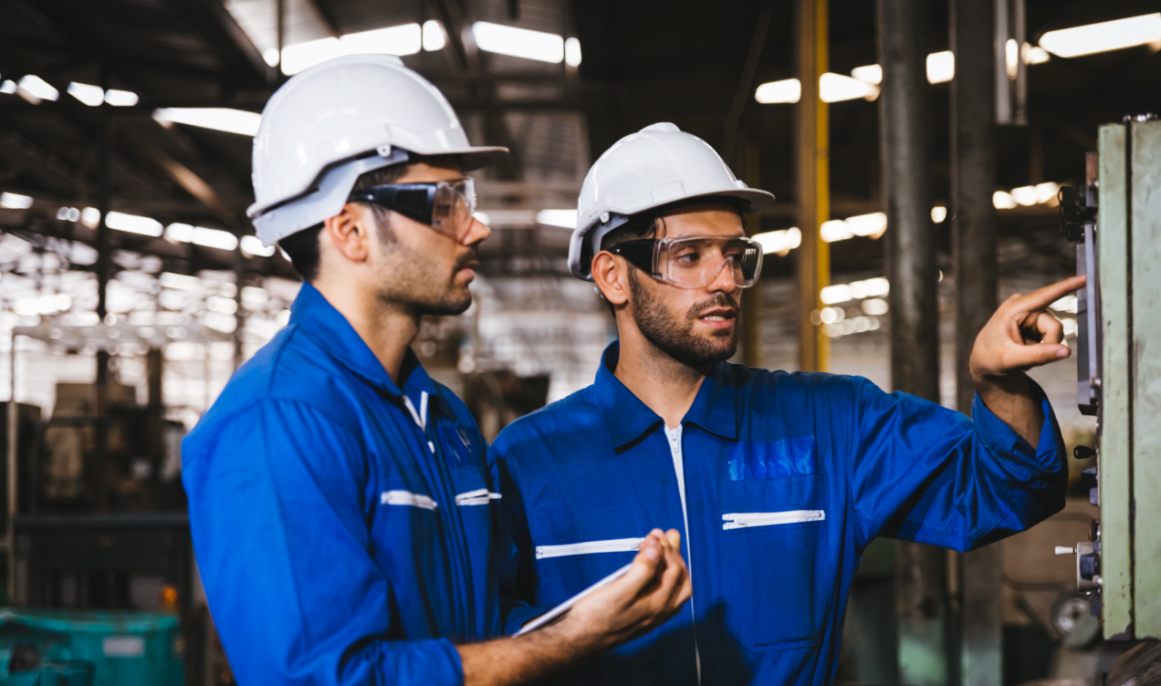 This image shows two industrial workers wearing blue coveralls, protective helmets, and safety glasses, collaborating in a factory or manufacturing setting. One worker is pointing at a control panel or machine, while the other is observing closely, indicating a discussion or inspection of the equipment. The industrial environment is characterized by metal structures and machinery in the background.