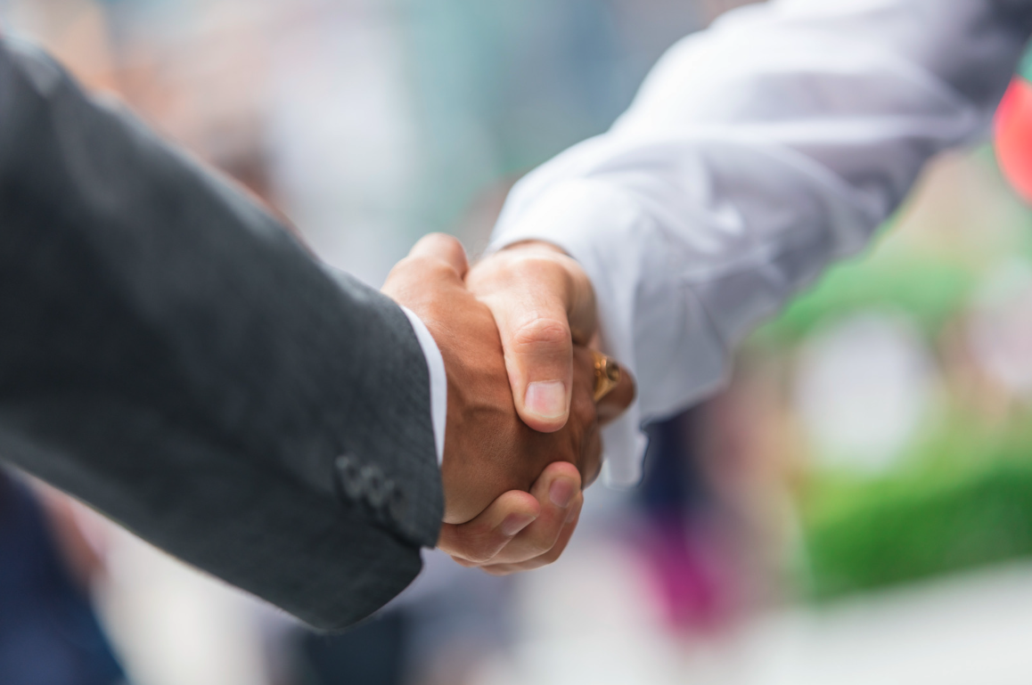 Close-up of two individuals in formal attire shaking hands, symbolizing agreement or partnership, with a blurred background suggesting an outdoor professional setting.