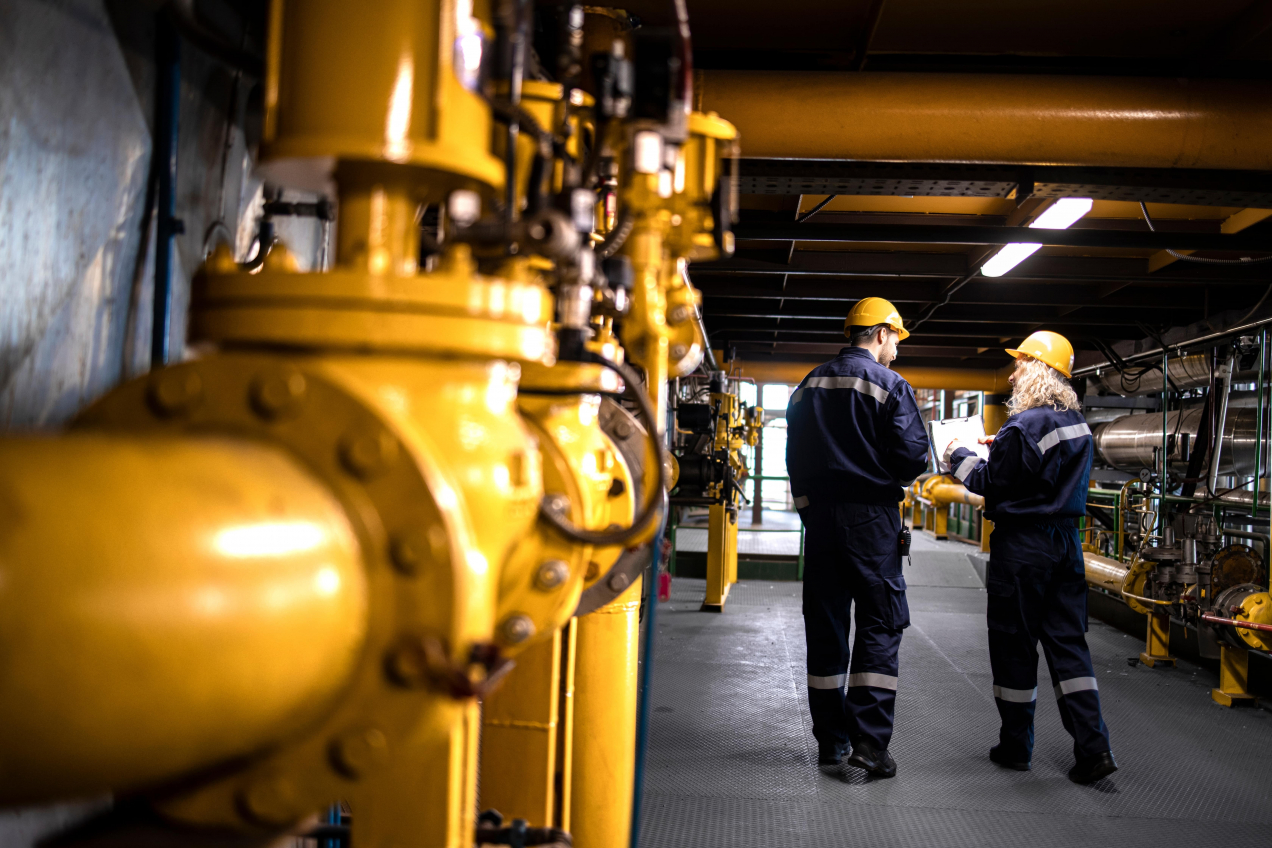 Two industrial workers, wearing yellow hard hats and navy-blue protective uniforms with reflective stripes, stand in a large, well-lit industrial facility. They are discussing information on a clipboard while surrounded by yellow pipelines and machinery. The environment appears clean and organized, with a focus on safety and communication.