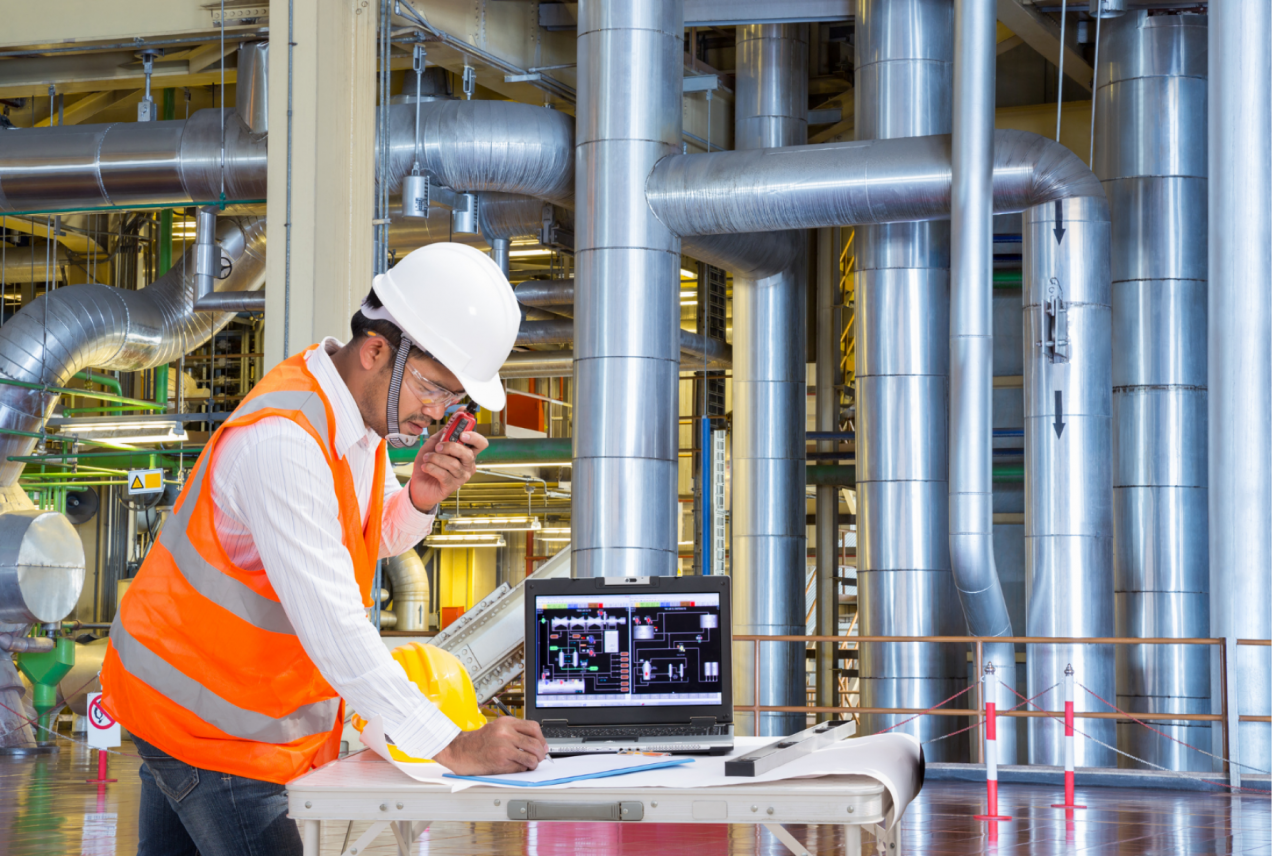 A male engineer wearing a hard hat, safety glasses, and a bright orange safety vest stands at a table in a large industrial facility. He is speaking into a radio while reviewing schematics and making notes on paper. A laptop on the table displays complex technical diagrams related to the facility's operations. In the background, large metallic pipes and equipment can be seen, indicating a busy manufacturing or processing environment.